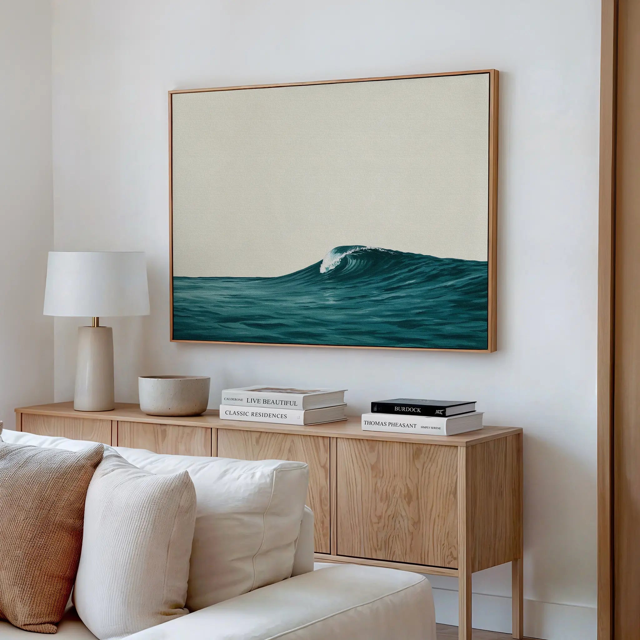 A modern living room features a wooden sideboard with books, a lamp, and a ceramic bowl. Above it hangs the Oceanside Crest Canvas Art, its ocean wave design complementing the white sofa and coastal decor.