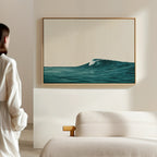 A person in a white outfit admires the Oceanside Crest Canvas Art—a framed blue ocean wave—hanging on a beige wall above a white sofa in a bright, minimal room, creating serene coastal living room decor.