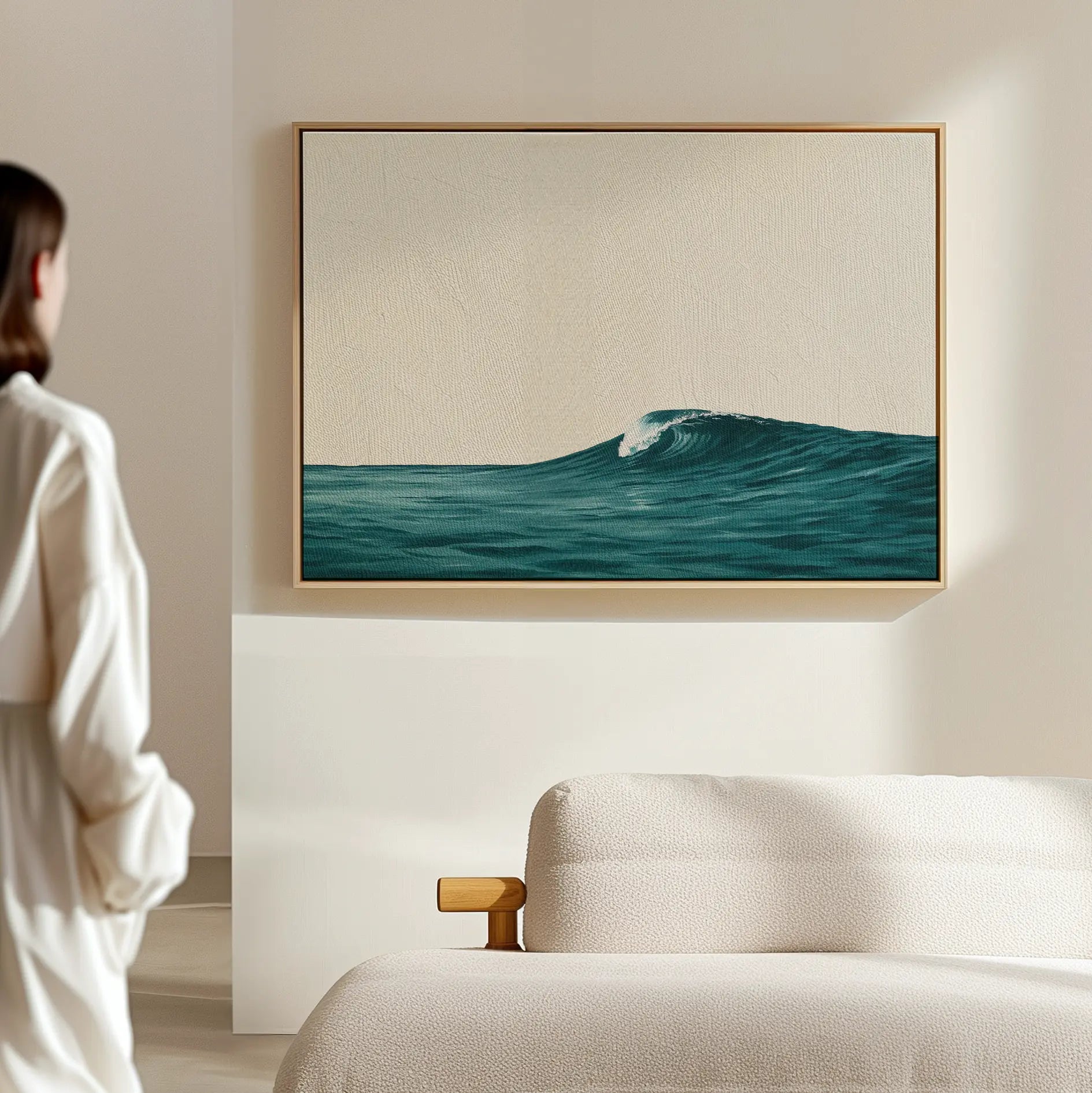 A person in a white outfit admires the Oceanside Crest Canvas Art—a framed blue ocean wave—hanging on a beige wall above a white sofa in a bright, minimal room, creating serene coastal living room decor.