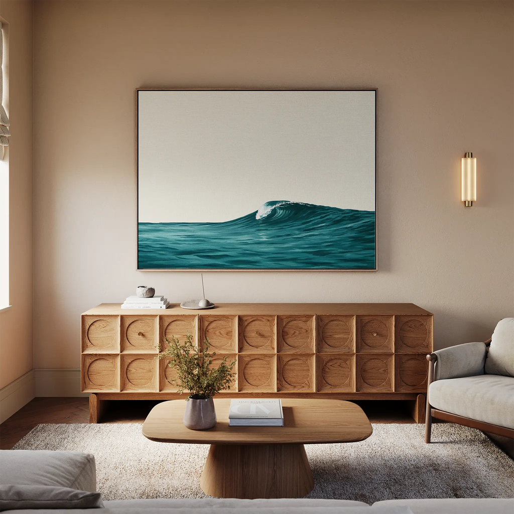 A modern living room with a wooden sideboard, low coffee table, light sofa, and coastal decor. Above the sideboard hangs the Oceanside Crest Canvas Art featuring a wave. A small vase of flowers decorates the coffee table.