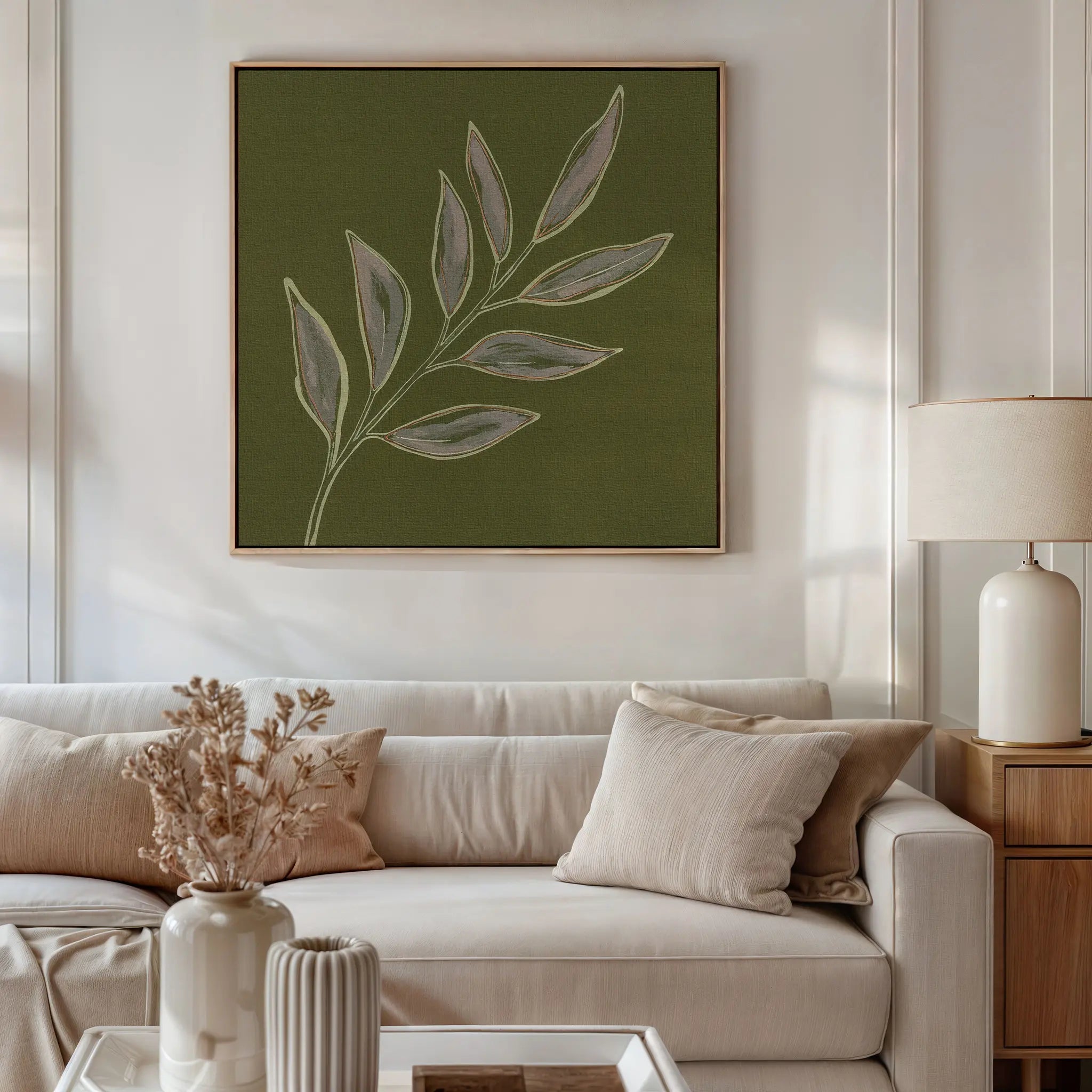 A modern living room featuring a beige sofa, cream pillows, a wooden side table with a white lamp, and the Olive Branch Canvas Art. A vase with dried flowers sits on a ribbed coffee table for refined decor.