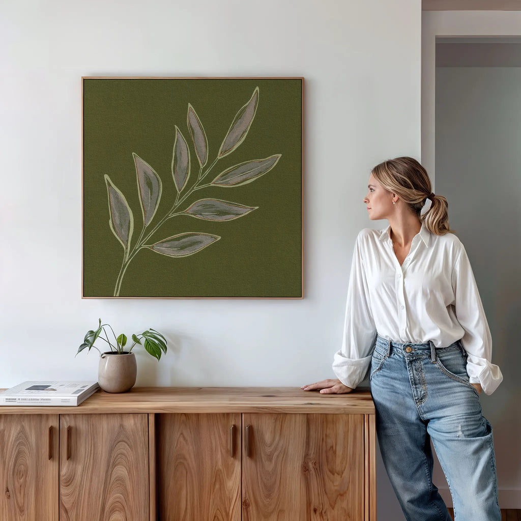 A woman in a white shirt and jeans leans against a wooden cabinet, admiring the Olive Branch Canvas Art above her. A potted plant and books rest on the cabinet, creating a calm, minimalistic atmosphere for modern living rooms.