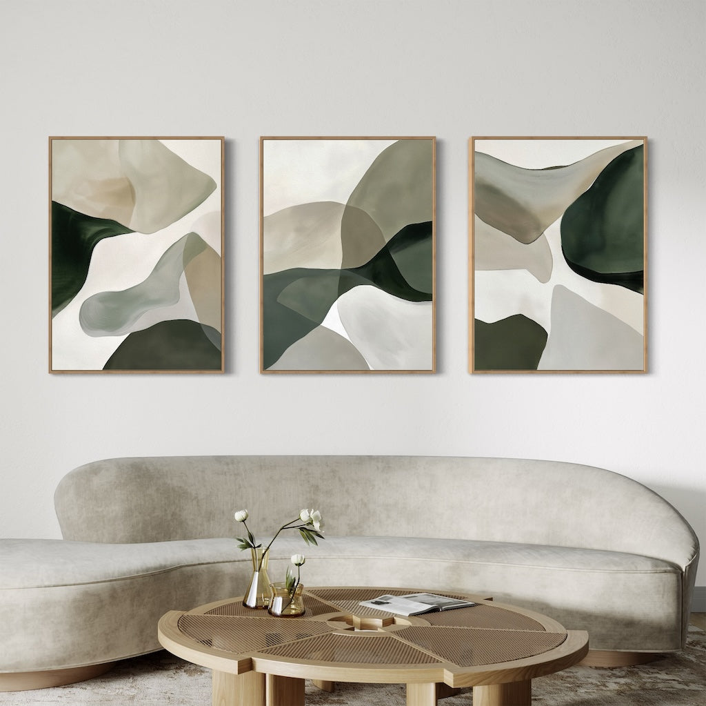 A modern living room features a curved beige sofa, round wooden coffee table, and the Organic Flow Set of 3 abstract green and beige paintings above the sofa. A small vase with white flowers completes this contemporary space.