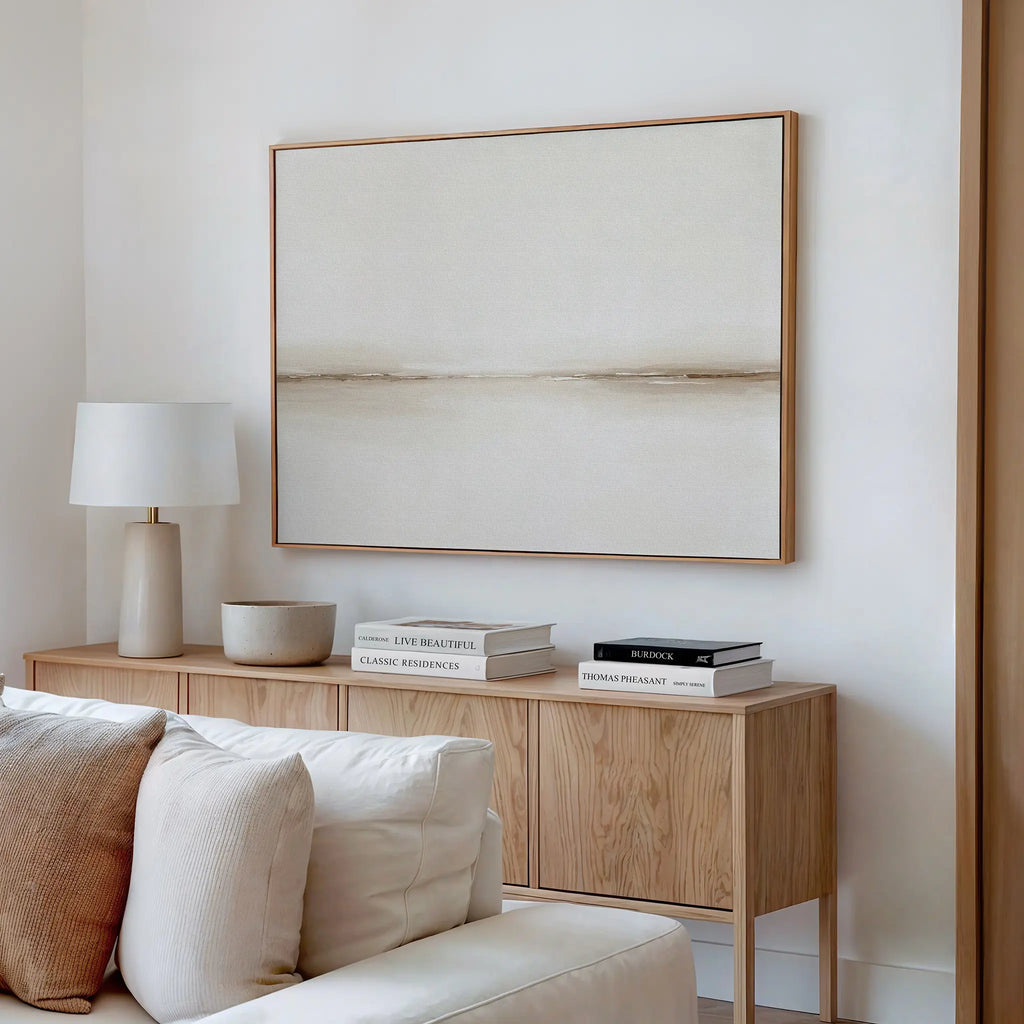 A minimalist living room with a wooden sideboard, neutral decor, books, a white table lamp, a bowl, and the Pale Desert Canvas Art in beige tones above a light-colored sofa—perfect for stylish living room decor.
