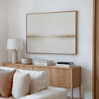 A minimalist living room with a wooden sideboard, neutral decor, books, a white table lamp, a bowl, and the Pale Desert Canvas Art in beige tones above a light-colored sofa—perfect for stylish living room decor.