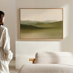 A person stands in a minimal, light-filled room, admiring the Pastoral Peace Canvas Art depicting rolling green hills displayed above a beige sofa.