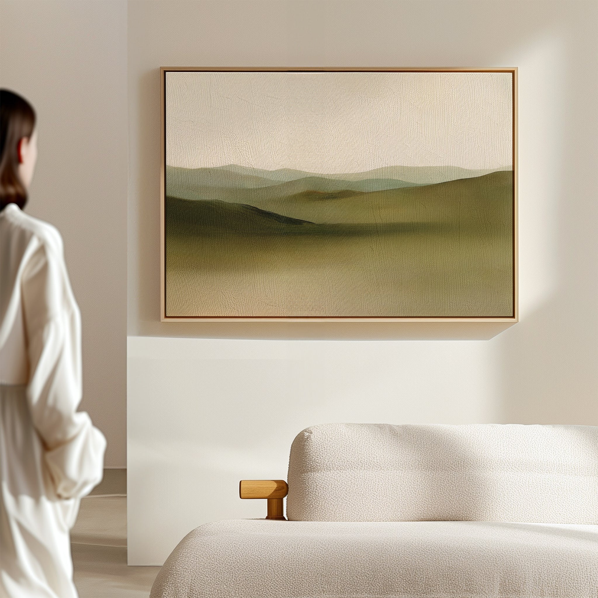 A person stands in a minimal, light-filled room, admiring the Pastoral Peace Canvas Art depicting rolling green hills displayed above a beige sofa.