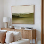 A modern living room with a light wood sideboard, books, a white lamp, and beige couch features minimalist decor and serene wall art—the Pastoral Peace Canvas Art, a large abstract landscape painting with green hills—for an inviting touch.