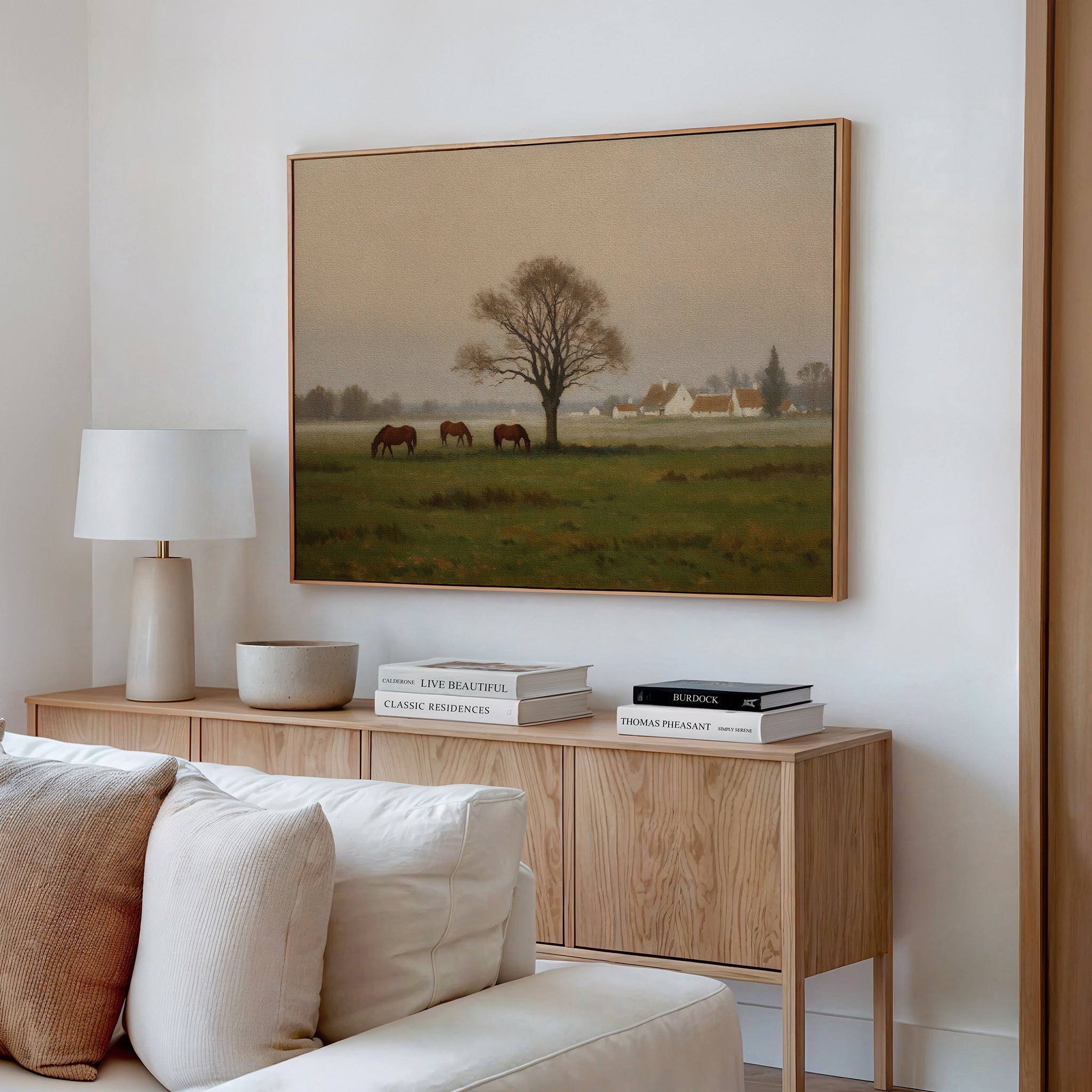 A cozy living room showcases a light wood sideboard with books, a lamp, and decor. Above it hangs the Pasture Morning Canvas Art, depicting horses grazing in a misty field. A neutral pillow sofa completes the rural vibe.