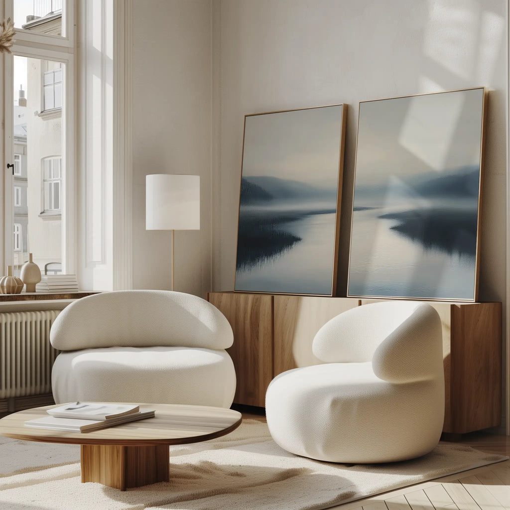 A modern living room features two rounded white armchairs, a low wooden coffee table, tranquil decor, and the Peaceful Waters Set of 2 landscape wall art leaning against a wooden cabinet. Soft natural light fills the space.