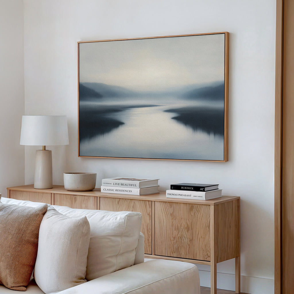 A serene living room features a wooden sideboard, white lamp, stacked books, and the "Peaceful Waters Canvas Art – Horizontal," a calming landscape above a cream-colored sofa.