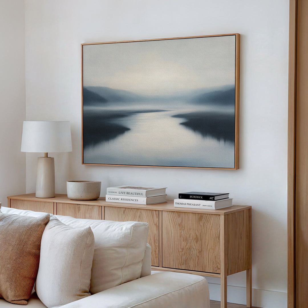 A serene living room features a wooden sideboard, white lamp, stacked books, and the "Peaceful Waters Canvas Art – Horizontal," a calming landscape above a cream-colored sofa.