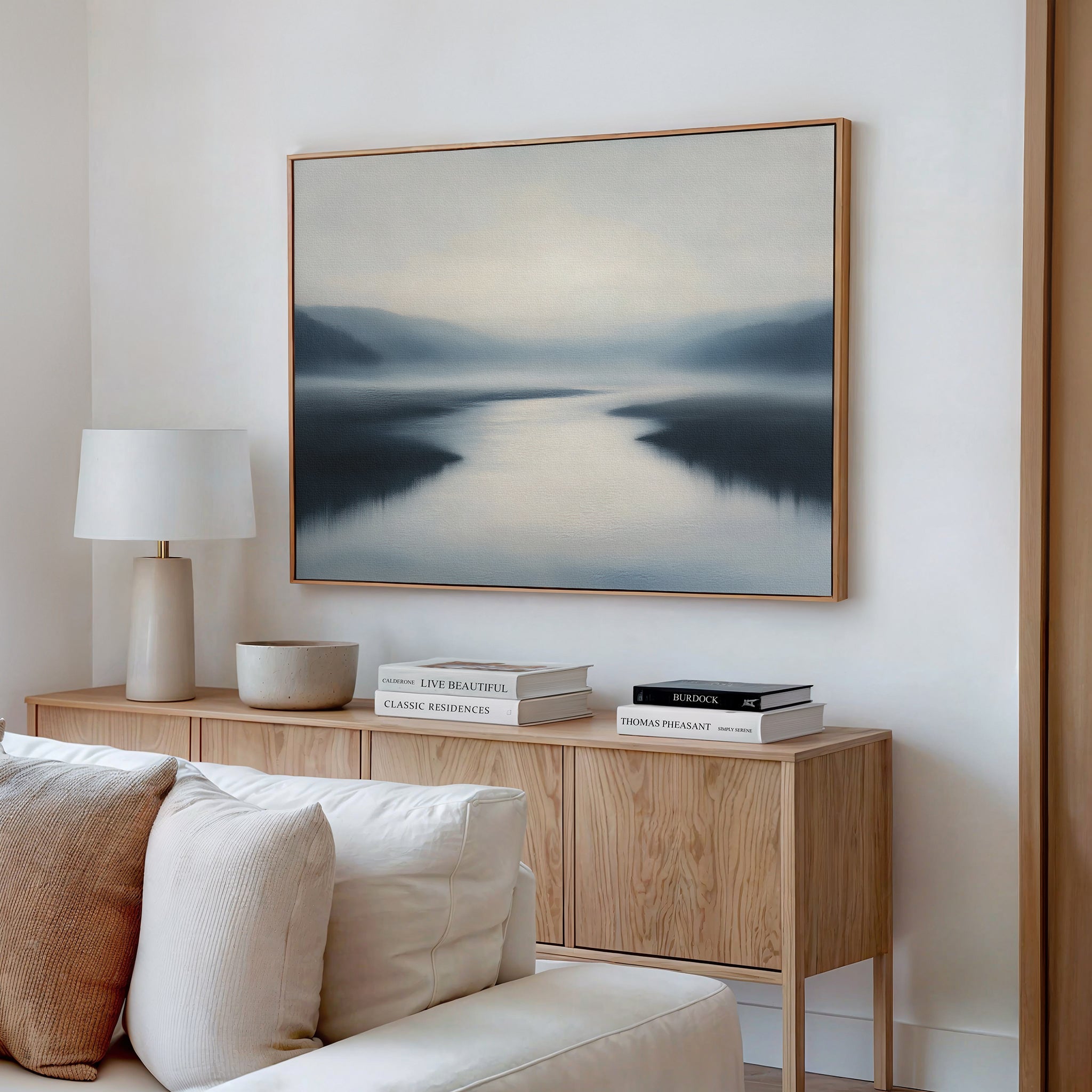 A serene living room features a wooden sideboard, white lamp, stacked books, and the "Peaceful Waters Canvas Art – Horizontal," a calming landscape above a cream-colored sofa.