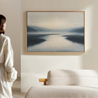 A person in light attire stands in a modern living room, admiring the "Peaceful Waters Canvas Art – Horizontal," a calming landscape painting of serene, misty waters and distant hills, hanging above the beige sofa.