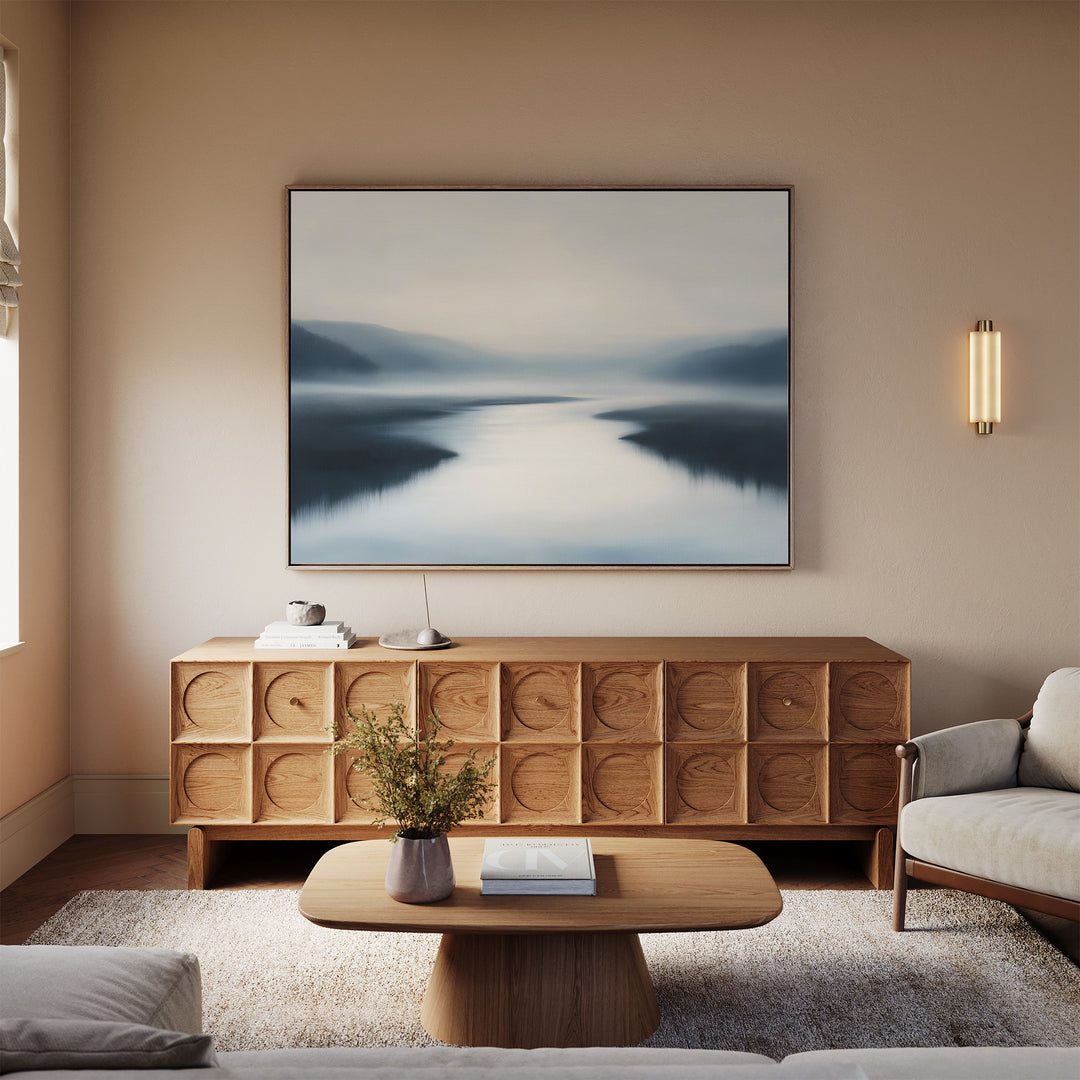 A modern living room with neutral tones features a wooden sideboard, low coffee table with flowers, plush rug, light sofa, and the Peaceful Waters Canvas Art – Horizontal above the sideboard for tranquil decor.