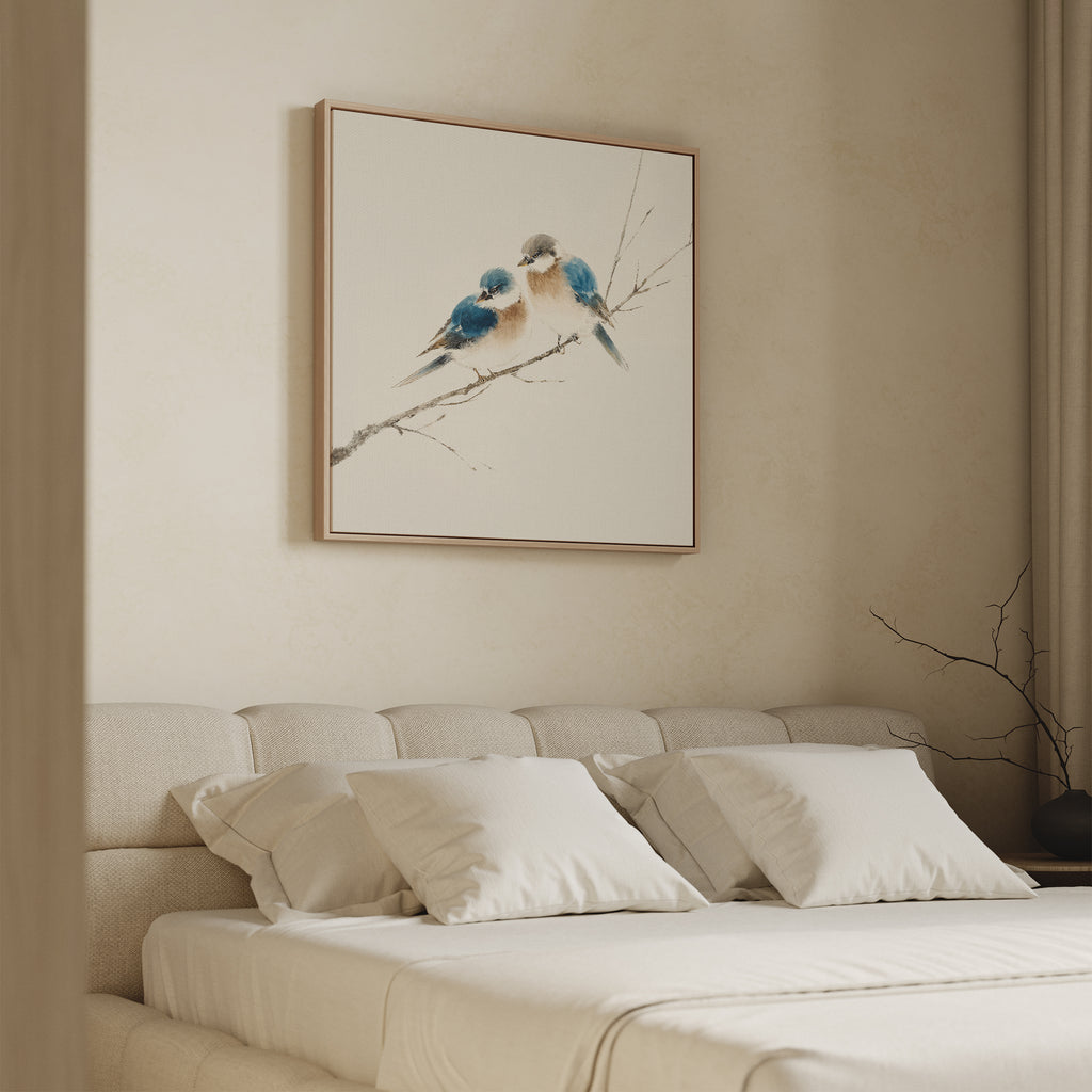 A minimalist bedroom with a beige upholstered bed, white bedding, and four pillows features Perched Companions Canvas Art—showing two blue birds on a branch—above the bed, while a bare branch decorates the bedside table.