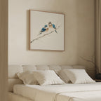 A minimalist bedroom with a beige upholstered bed, white bedding, and four pillows features Perched Companions Canvas Art—showing two blue birds on a branch—above the bed, while a bare branch decorates the bedside table.