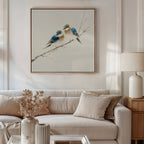 A neutral living room features a white sofa, beige cushions, and a side table with a lamp. Above the couch is the Perched Companions Canvas Art—framed bluebirds on a branch. A vase of dried flowers sits on the coffee table.
