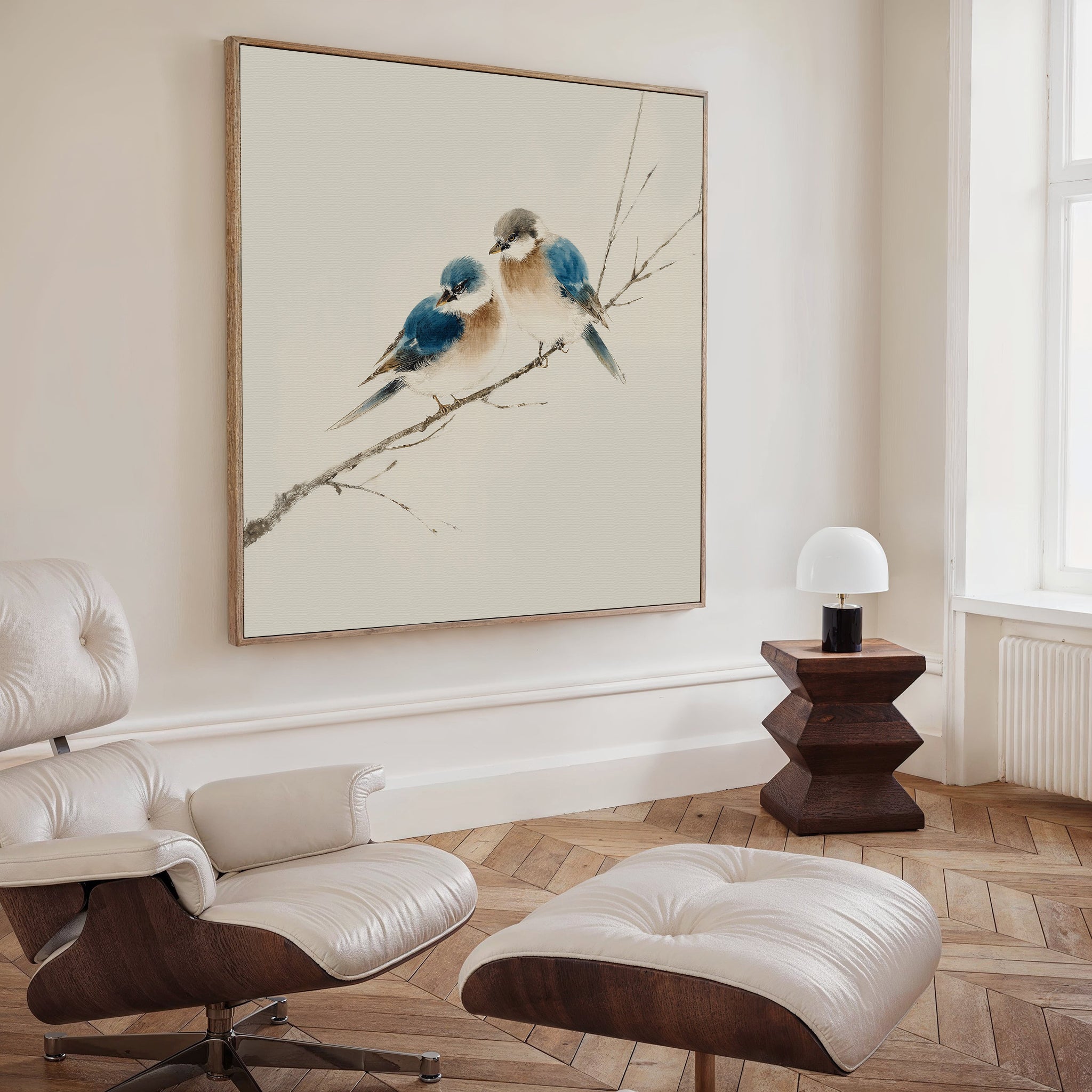 A cozy living room featuring a modern white lounge chair with ottoman, a wooden side table and lamp, and Perched Companions Canvas Art as striking Nordic wall decor. Sunlight pours in through a tall window.