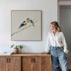 A woman in a white shirt and jeans leans on a wooden cabinet topped with a small plant and books, gazing at the Perched Companions Canvas Art—a Nordic wall decor print of two bluebirds resting on a branch.