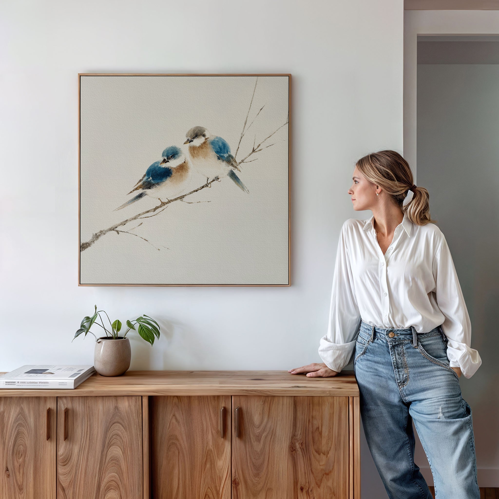 A woman in a white shirt and jeans leans on a wooden cabinet topped with a small plant and books, gazing at the Perched Companions Canvas Art—a Nordic wall decor print of two bluebirds resting on a branch.