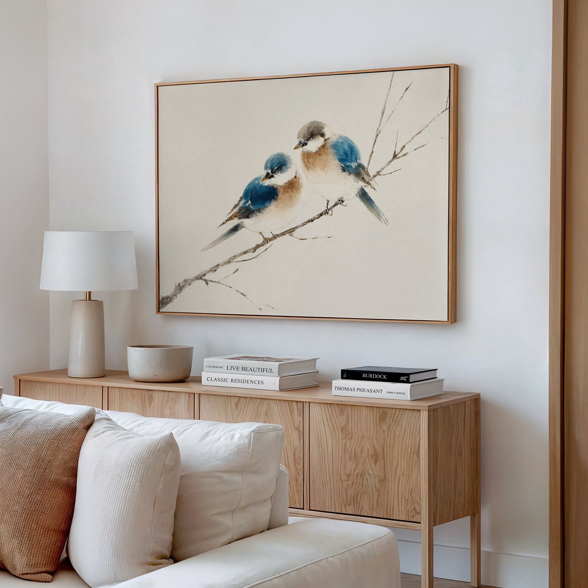 A minimalist living room with a light wood sideboard, neutral decor, and the Perched Companions Canvas Art – Horizontal featuring two bluebirds above. A white lamp and stacked books complete the cozy look.