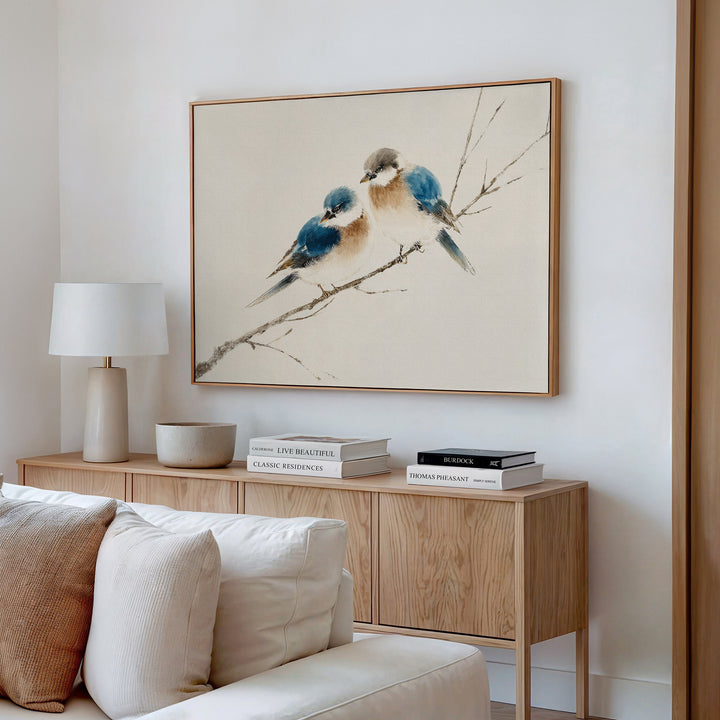 A minimalist living room with a light wood sideboard, neutral decor, and the Perched Companions Canvas Art – Horizontal featuring two bluebirds above. A white lamp and stacked books complete the cozy look.