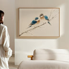 In a softly lit, minimal room, a person admires the Perched Companions Canvas Art – Horizontal featuring two blue and brown birds on a branch above a beige bed—a perfect accent for cozy home decor.