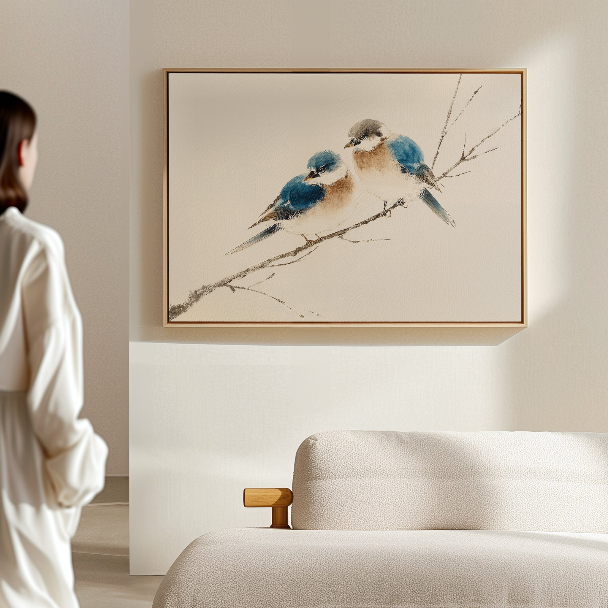 In a softly lit, minimal room, a person admires the Perched Companions Canvas Art – Horizontal featuring two blue and brown birds on a branch above a beige bed—a perfect accent for cozy home decor.