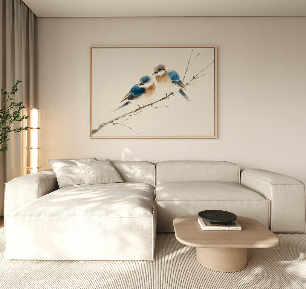 A modern living room featuring a light beige sectional sofa, a low wooden coffee table, and the "Perched Companions Canvas Art – Horizontal" print of two bluebirds on a branch above the couch, with a tall plant in the corner for cozy decor.