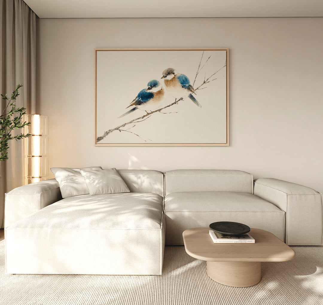 A modern living room featuring a light beige sectional sofa, a low wooden coffee table, and the "Perched Companions Canvas Art – Horizontal" print of two bluebirds on a branch above the couch, with a tall plant in the corner for cozy decor.