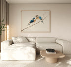A modern living room featuring a light beige sectional sofa, a low wooden coffee table, and the "Perched Companions Canvas Art – Horizontal" print of two bluebirds on a branch above the couch, with a tall plant in the corner for cozy decor.
