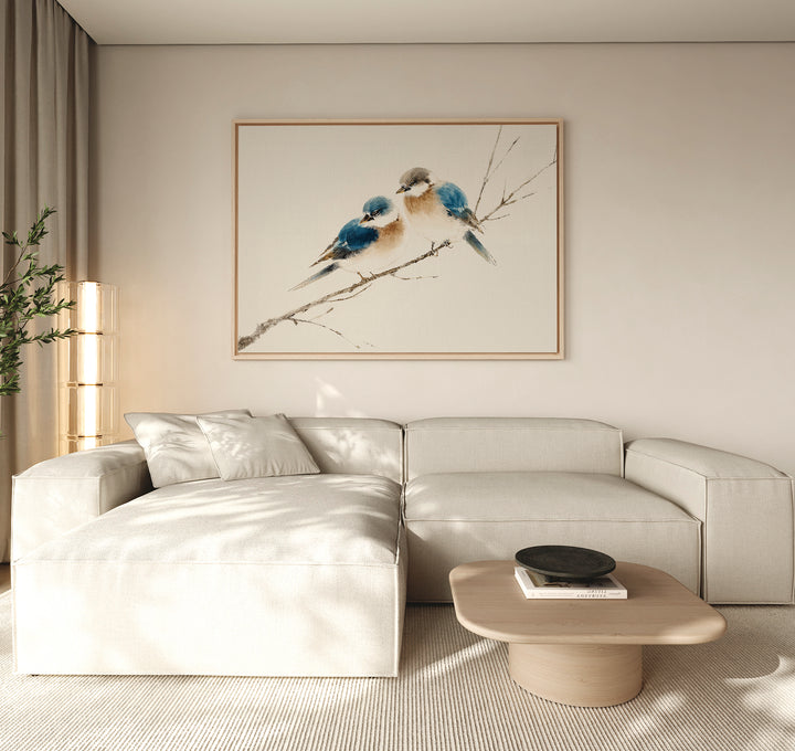 A modern living room featuring a light beige sectional sofa, a low wooden coffee table, and the "Perched Companions Canvas Art – Horizontal" print of two bluebirds on a branch above the couch, with a tall plant in the corner for cozy decor.