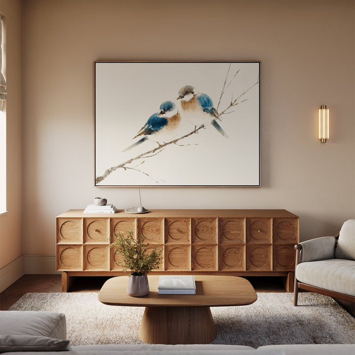 A cozy living room features a light wood sideboard, the "Perched Companions Canvas Art – Horizontal" wall print of two bluebirds on a branch, a coffee table with a vase and book, a beige sofa, and warm neutral walls with soft natural light.