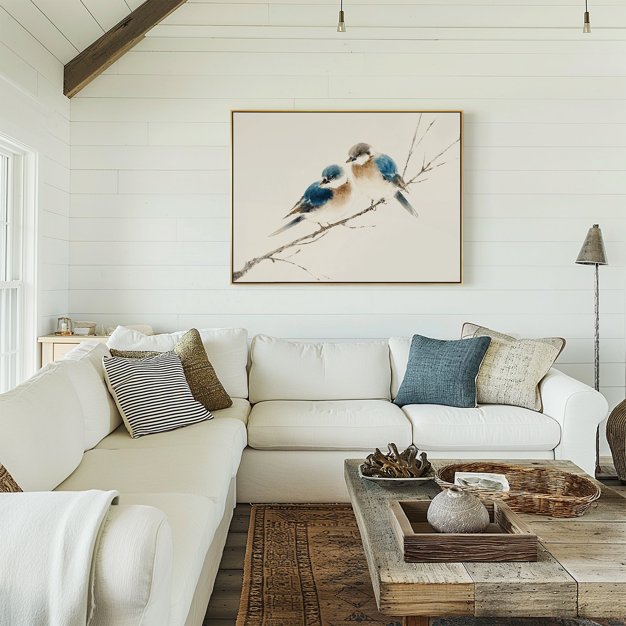 A bright, cozy living room with a white L-shaped sofa, neutral pillows, wooden coffee table, and the Perched Companions Canvas Art – Horizontal featuring two blue birds on a branch hanging on a white shiplap wall.