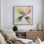 A modern living room featuring a neutral sofa, striped cushions, light wood cabinet, decorative accents, and the Petal Hues Canvas Art—a large abstract floral print in soft greens, yellows, and pinks—displayed on a white wall.