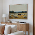 A modern living room with a light wood sideboard, books, lamp, and decorative bowl features Prairie Silence Canvas Art above—a bison in a grassy field with trees and mountains—bringing nature-inspired style to the white wall.