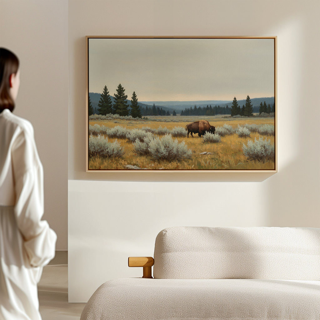 In a minimal, modern room, a person admires the Prairie Silence Canvas Art—a framed painting of a bison in grassy fields with pines and soft mountains—bringing nature-inspired décor to the serene space.