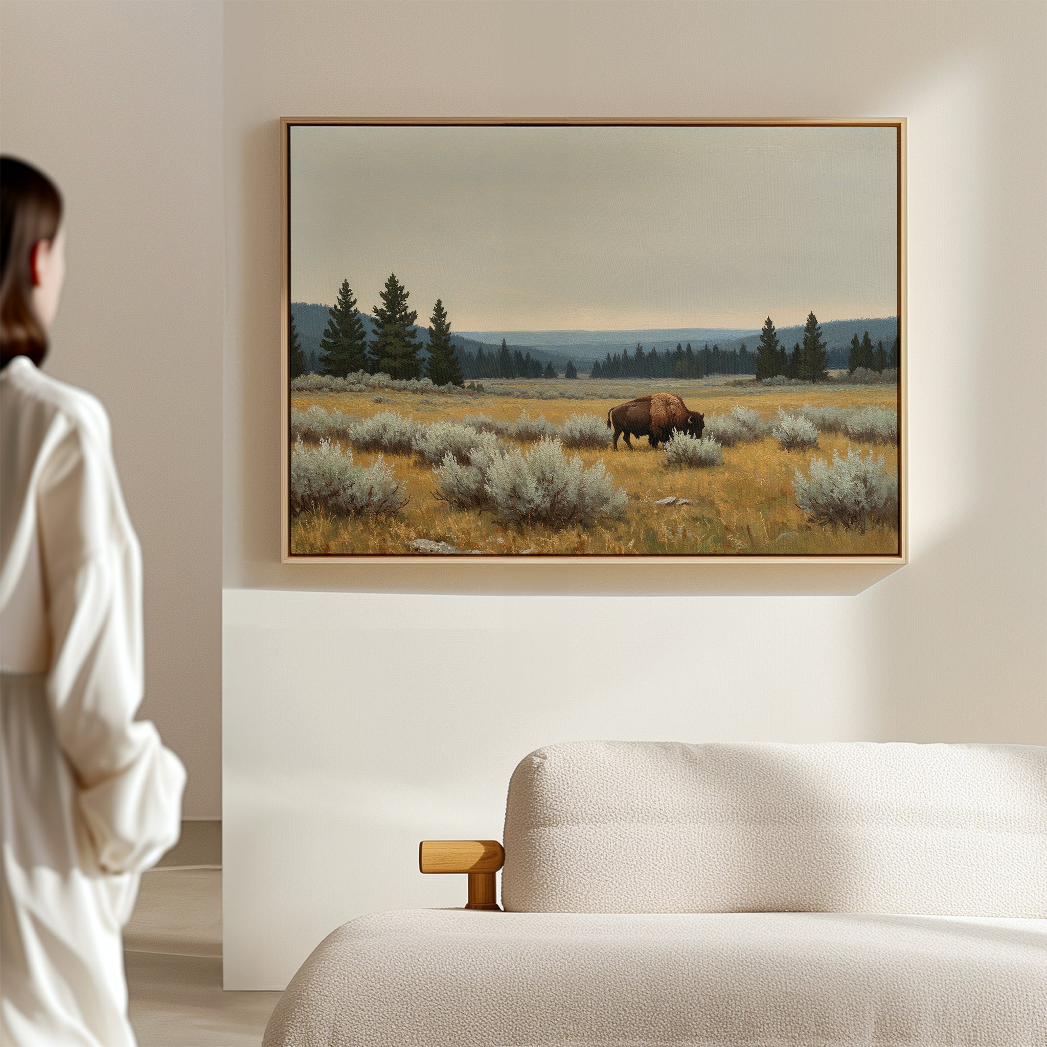 In a minimal, modern room, a person admires the Prairie Silence Canvas Art—a framed painting of a bison in grassy fields with pines and soft mountains—bringing nature-inspired décor to the serene space.