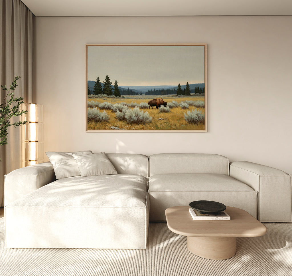 A modern living room features a beige sectional, a light wood coffee table, and the Prairie Silence Canvas Art—a large landscape of a bison in a grassy field—adding nature-inspired decor to the light wall.