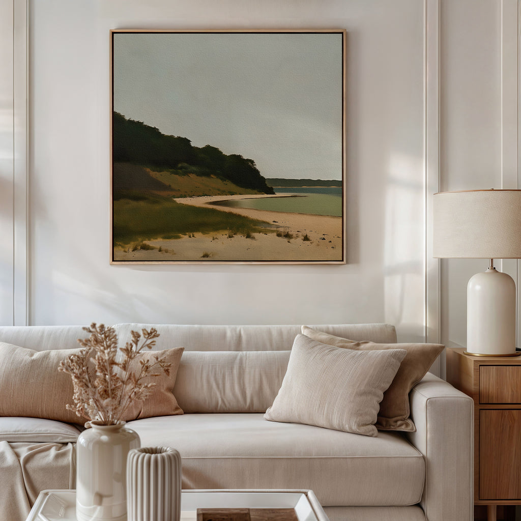 A minimalist living room features a white sofa, beige pillows, and a side table with a white lamp. Above the sofa, the Quiet Bend Canvas Art—large-scale wall decor depicting a beach landscape with greenery—adds chic style.