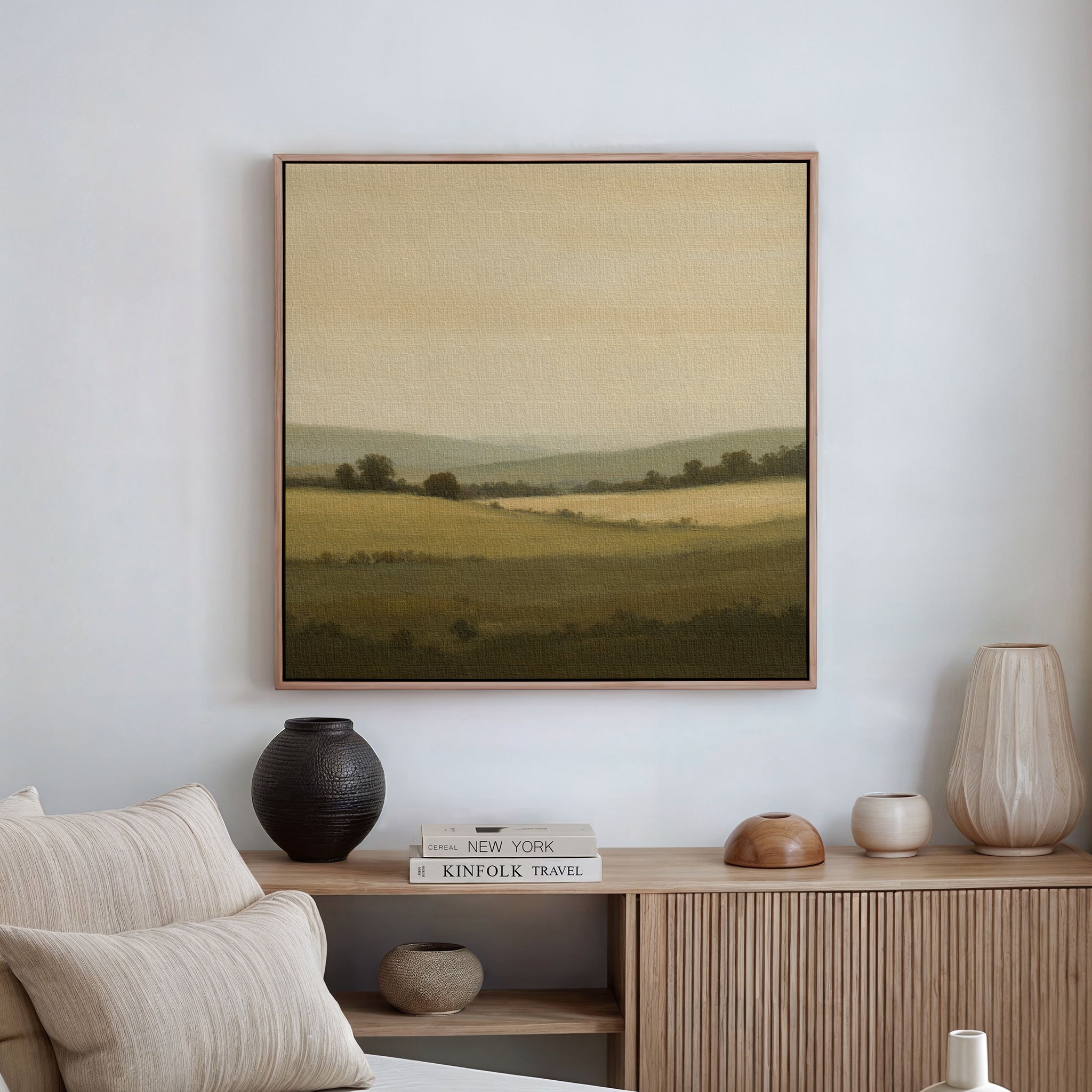A minimal living room features a beige sofa, neutral decor, and the Quiet Countryside Canvas Art depicting rolling hills and trees as minimalist wall art above a wooden sideboard with vases, a candle, and stacked books.