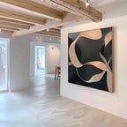 A modern, minimal art gallery with white walls displays the Quiet Division Abstract Canvas Art—featuring beige and black swirling shapes. Light wood beams and natural light enhance the elegant, contemporary space.