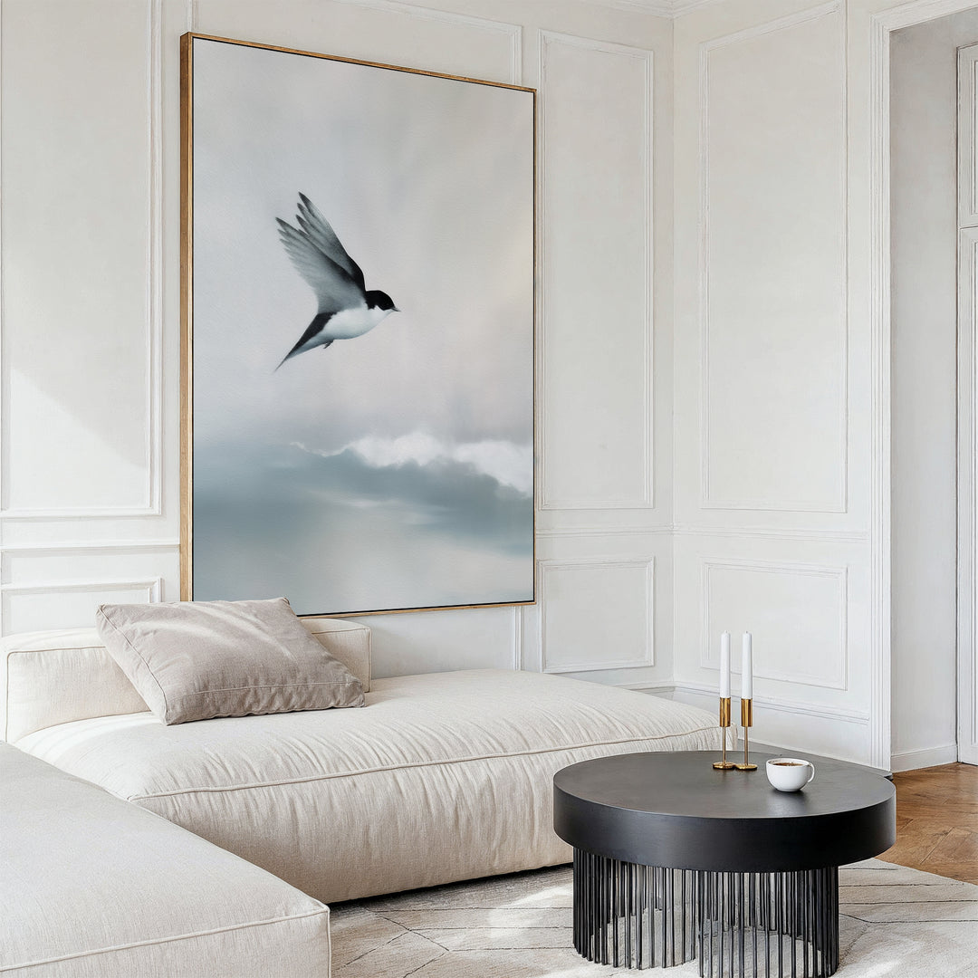 A modern living room with a cream-colored sectional sofa, black round coffee table, and Quiet Flight Canvas Art featuring a bird soaring over soft clouds creates striking minimalist wall decor.