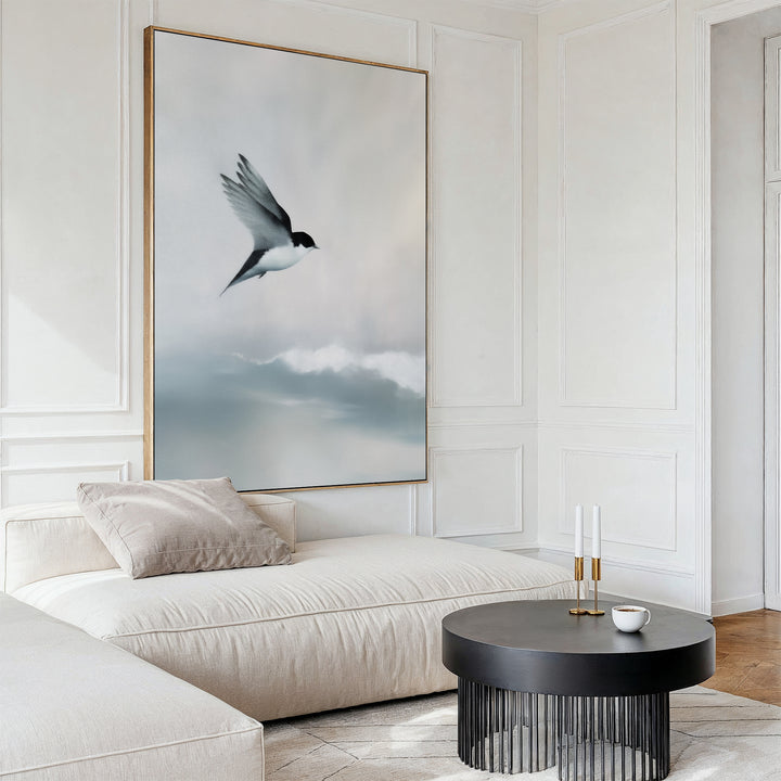 A modern living room with a cream-colored sectional sofa, black round coffee table, and Quiet Flight Canvas Art featuring a bird soaring over soft clouds creates striking minimalist wall decor.