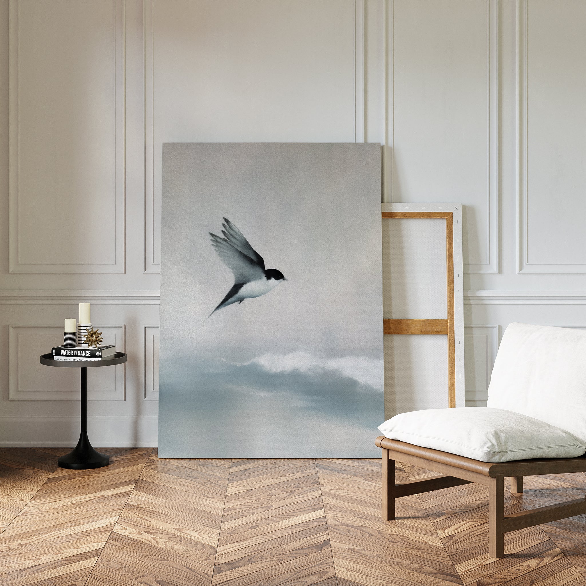 A minimalist Nordic Park-inspired room features the Quiet Flight Canvas Art of a flying bird, leaning against a white paneled wall beside a wood chair with cushion and a round table with candles and books on parquet flooring.