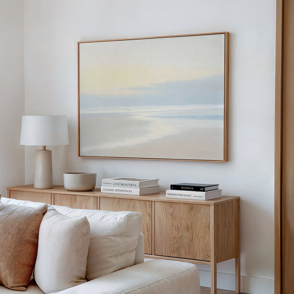 A minimal living room features a light wood sideboard, white table lamp, decorative bowl, stacked books, and the Quiet Shores Canvas Art – Horizontal displayed above a beige couch on a white wall.