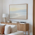 A minimal living room features a light wood sideboard, white table lamp, decorative bowl, stacked books, and the Quiet Shores Canvas Art – Horizontal displayed above a beige couch on a white wall.