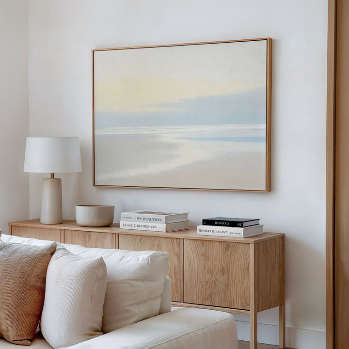 A minimal living room features a light wood sideboard, white table lamp, decorative bowl, stacked books, and the Quiet Shores Canvas Art – Horizontal displayed above a beige couch on a white wall.