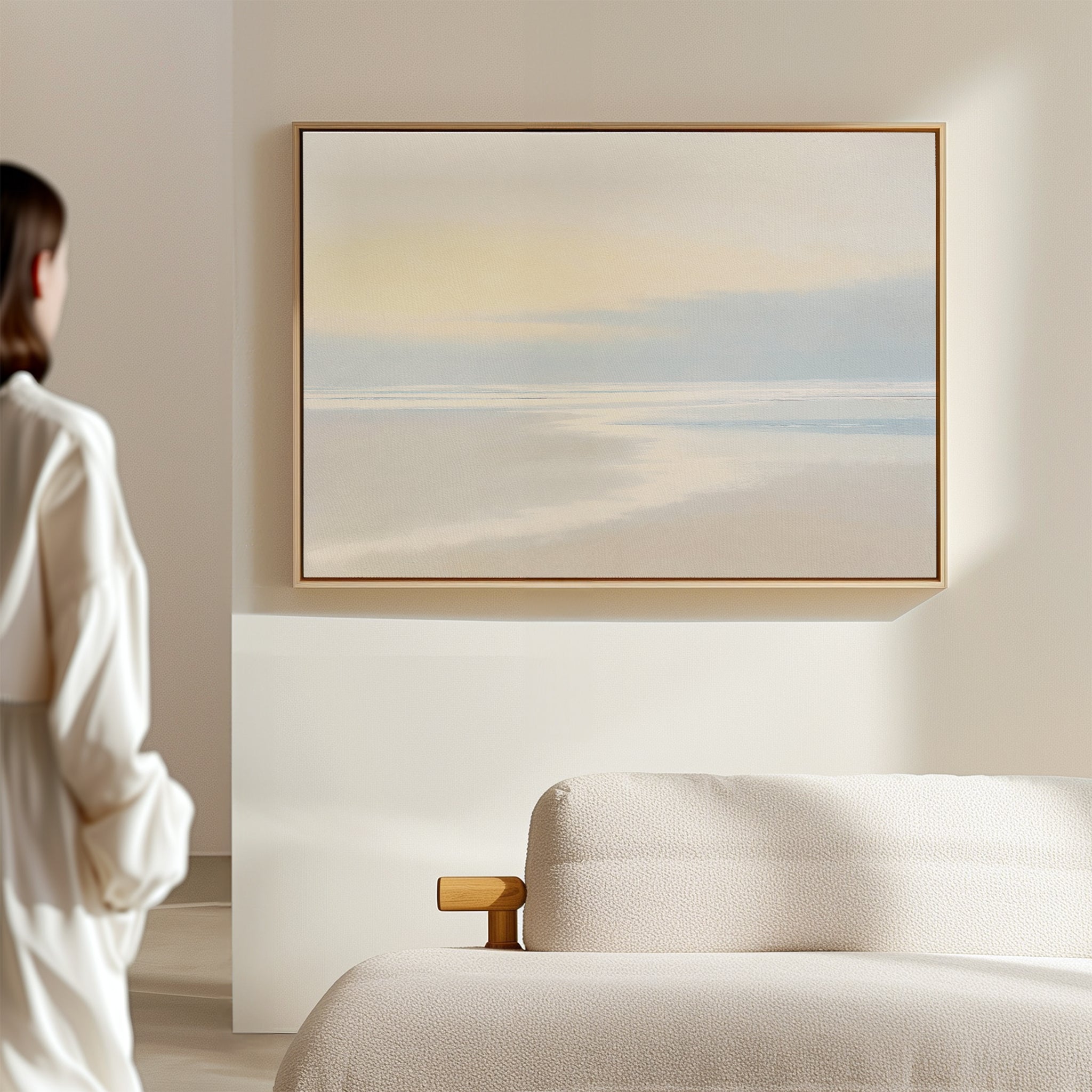 A person in white stands in a bright, minimalist room, admiring the Quiet Shores Canvas Art – Horizontal above a beige sofa—perfect for adding serene coastal decor to any space.